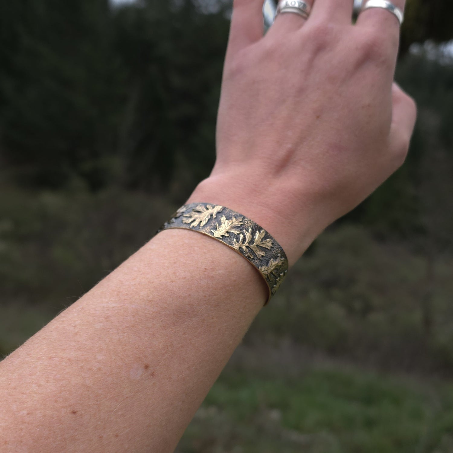 A hand reaching out with a background of dark leaves. The wrist of the hand is wearing a bracelet with golden colored oak leaves on it