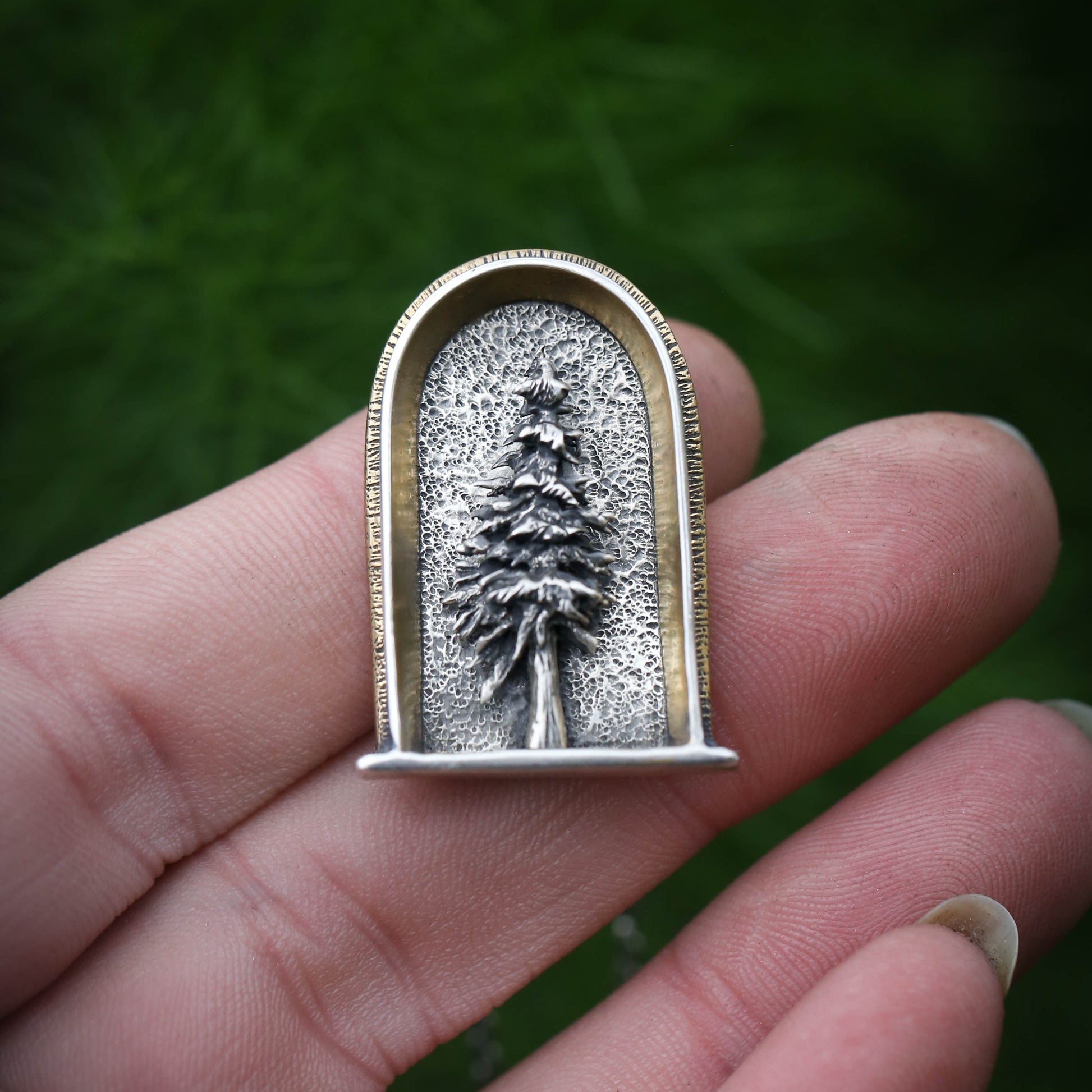 A sterling silver arched shrine shaped necklace featuring a three dimensional evergreen tree with a gold and brass border sits on a hand with a dark background