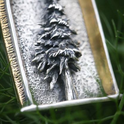 An up close picture of a sterling silver arched shrine shaped necklace featuring a three dimensional evergreen tree with a gold and brass border on a dark background