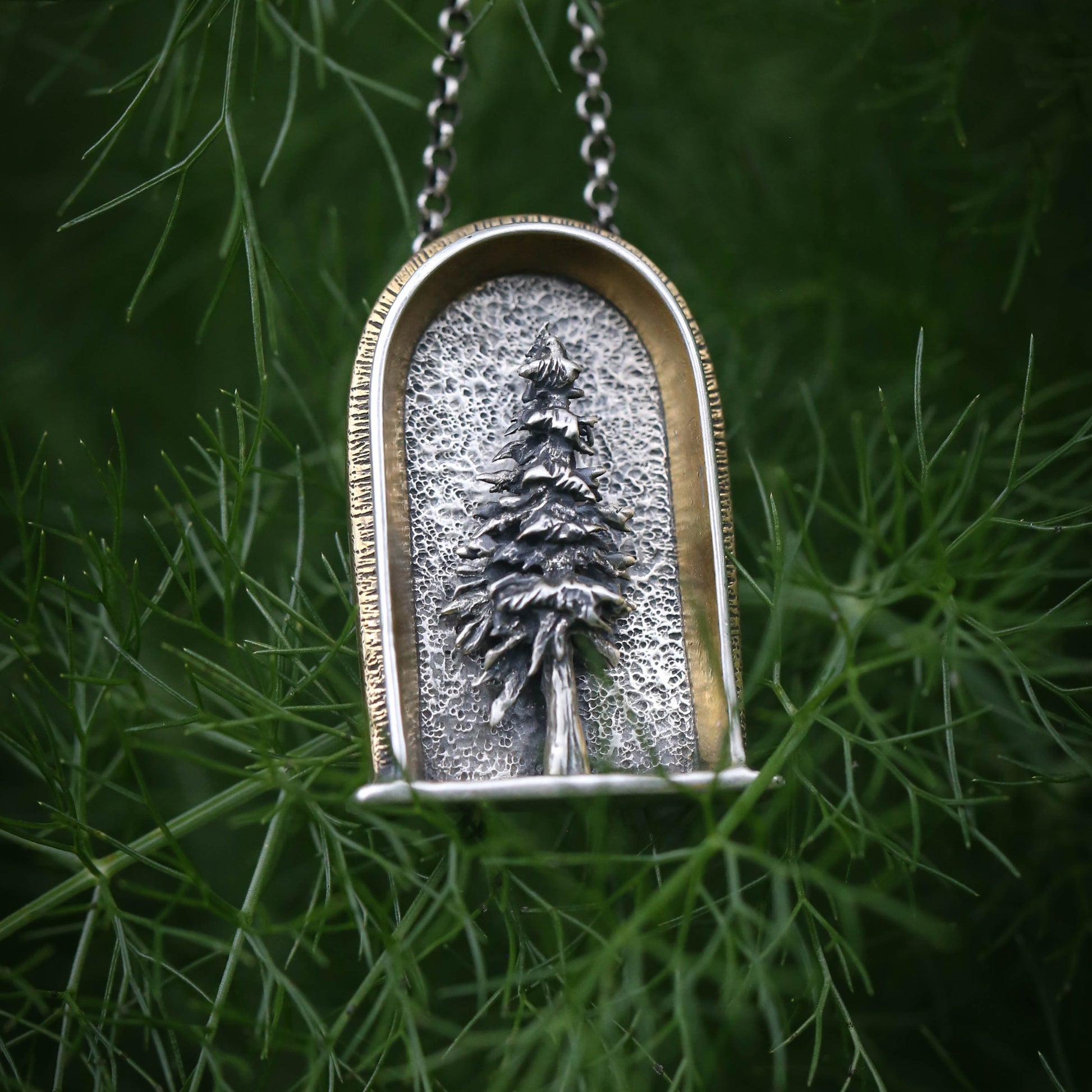 A sterling silver arched shrine shaped necklace featuring a three dimensional evergreen tree with a gold and brass border on a dark natural background