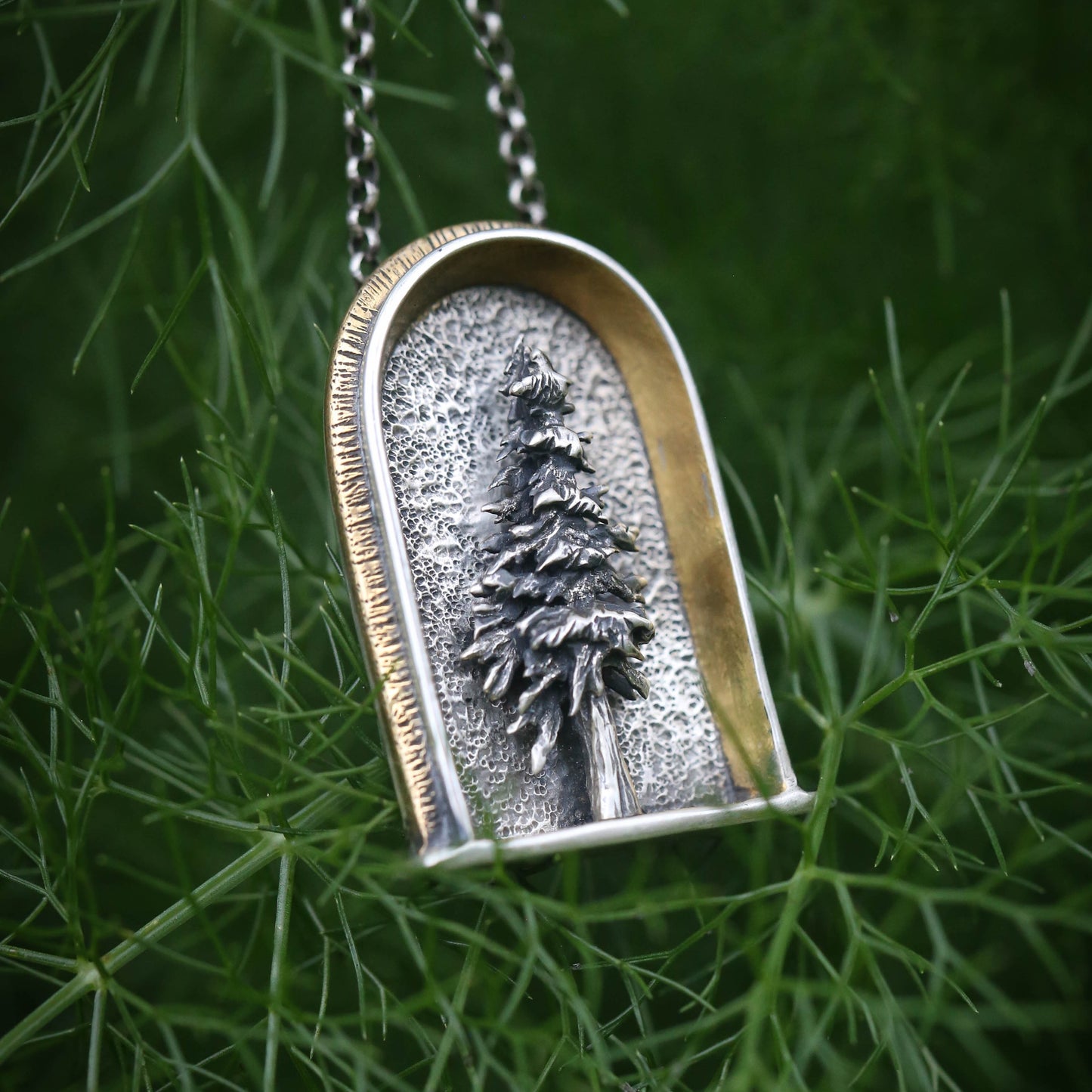 A sterling silver arched shrine shaped necklace featuring a three dimensional evergreen tree with a gold and brass border on a dark natural background