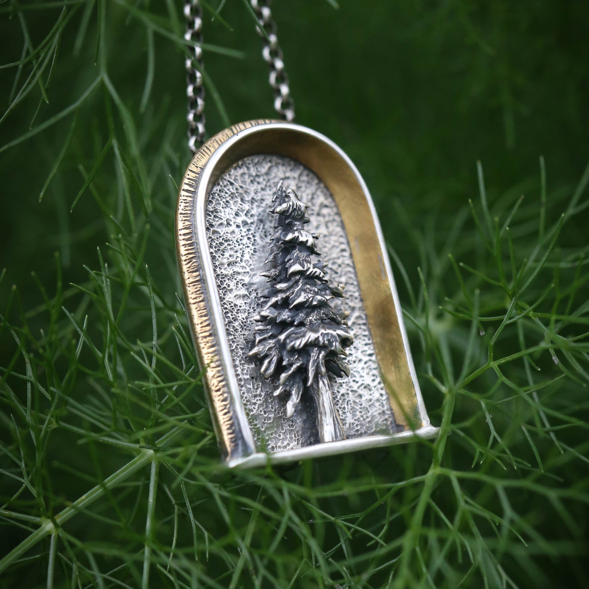 A sterling silver arched shrine shaped necklace featuring a three dimensional evergreen tree with a gold and brass border on a dark natural background