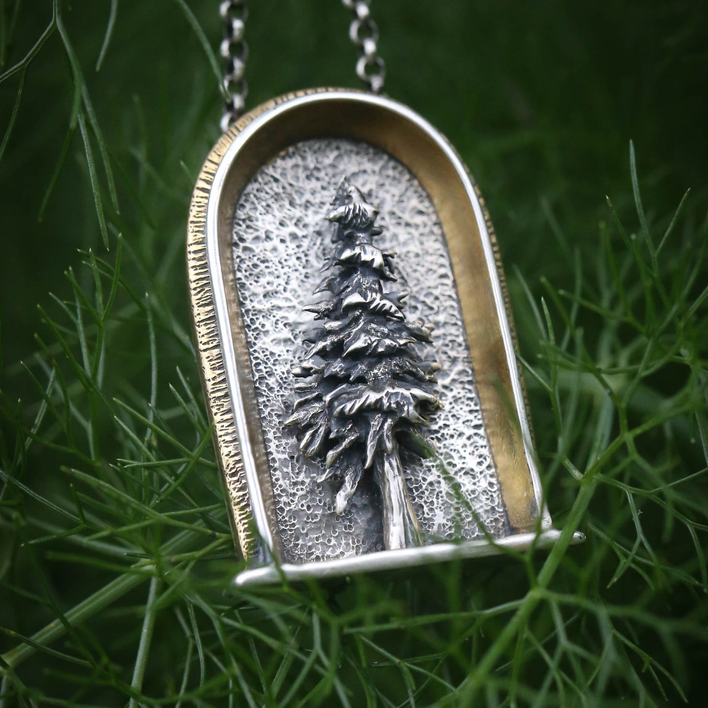 A sterling silver arched shrine shaped necklace featuring a three dimensional evergreen tree with a gold and brass border on a dark natural background