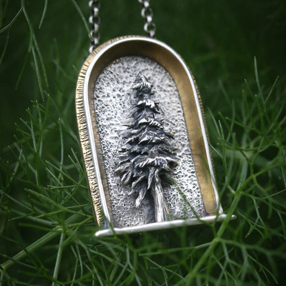A sterling silver arched shrine shaped necklace featuring a three dimensional evergreen tree with a gold and brass border on a dark natural background