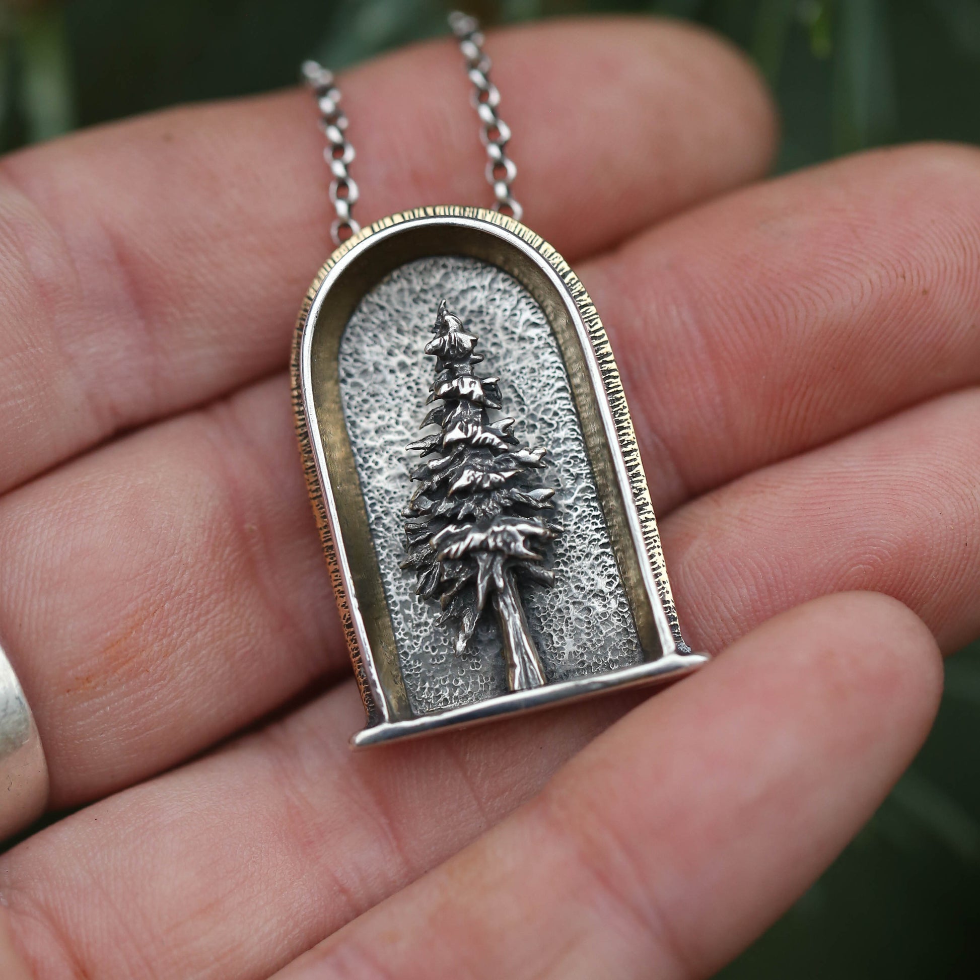 A sterling silver, 24k gold and brass necklace featuring a three-dimensional evergreen tree design, held in a person's hand.