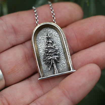 A sterling silver, 24k gold and brass necklace featuring a three-dimensional evergreen tree design, held in a person's hand.