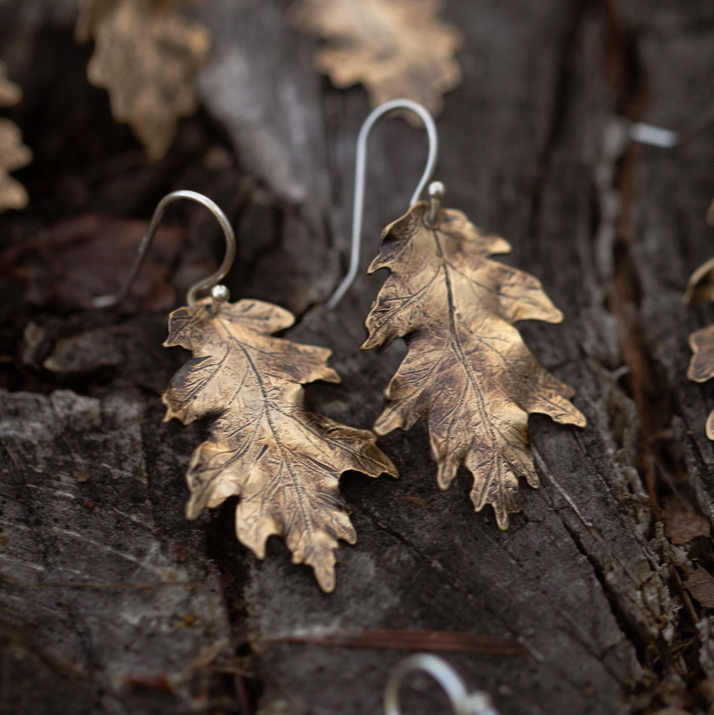 Brass gold colored oak leaf-shaped earrings on a textured wooden surface.