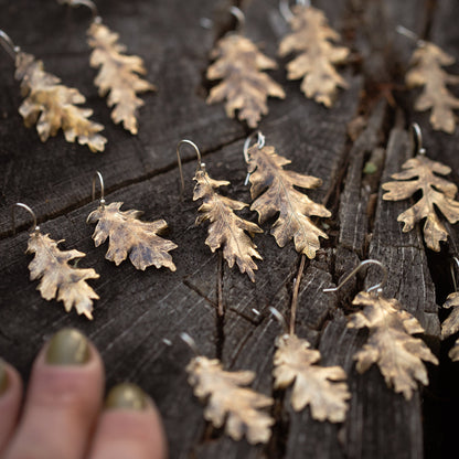 Pair of brass oak leaf-shaped earrings on a wooden surface with a hand near them surrounded by more pairs of oak leaf earrings