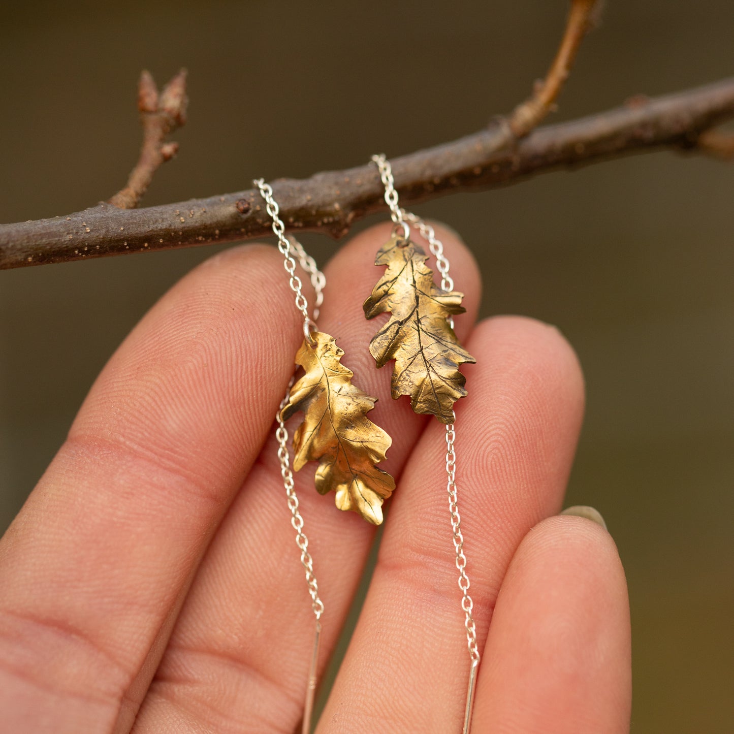 Brass oak leaf-shaped ear threader earrings held against a blurred natural background
