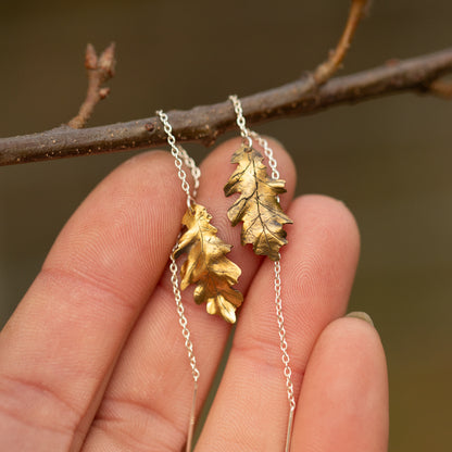 Brass oak leaf-shaped ear threader earrings held against a blurred natural background