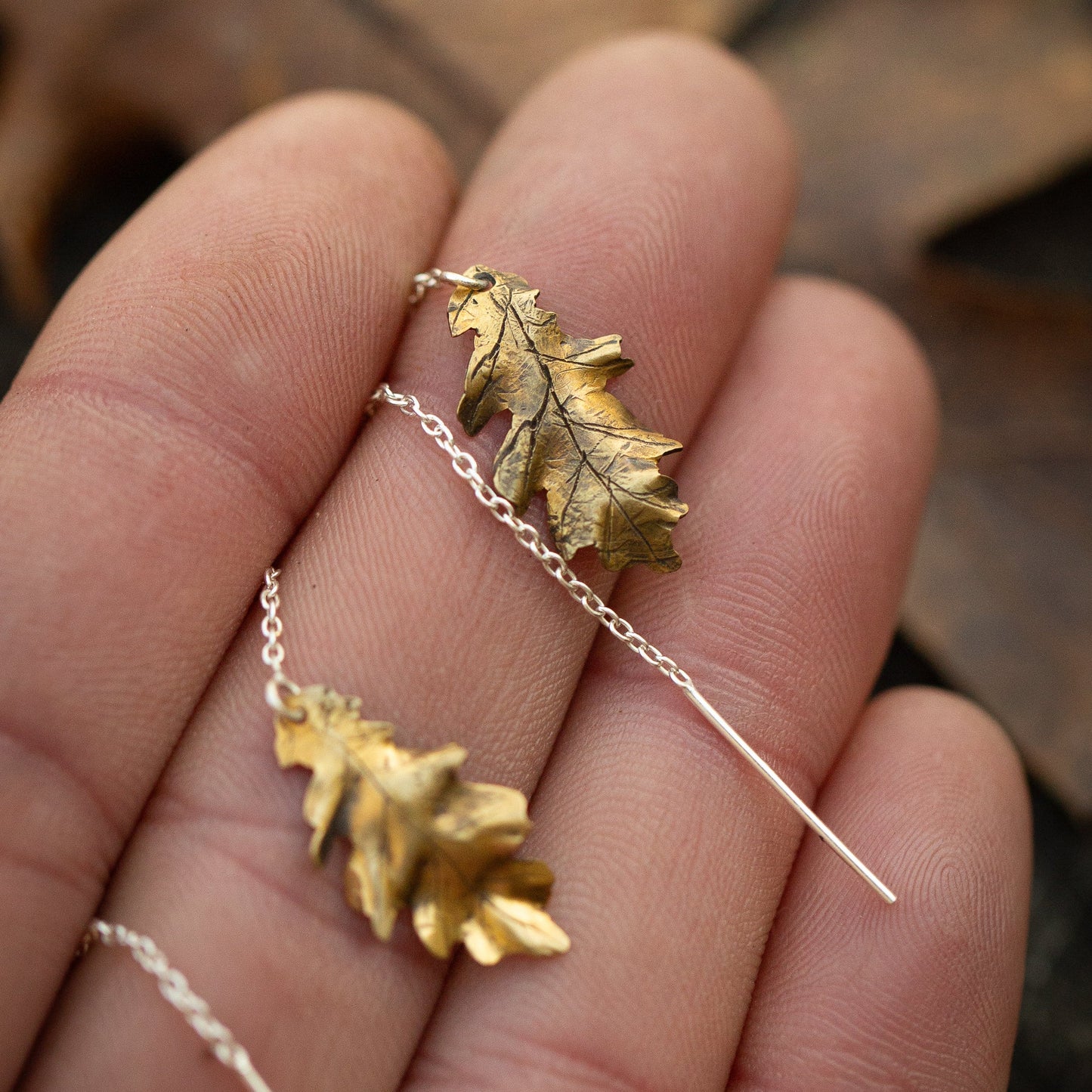 Brass gold colored oak leaf ear threader earring on a hand with a blurred natural background