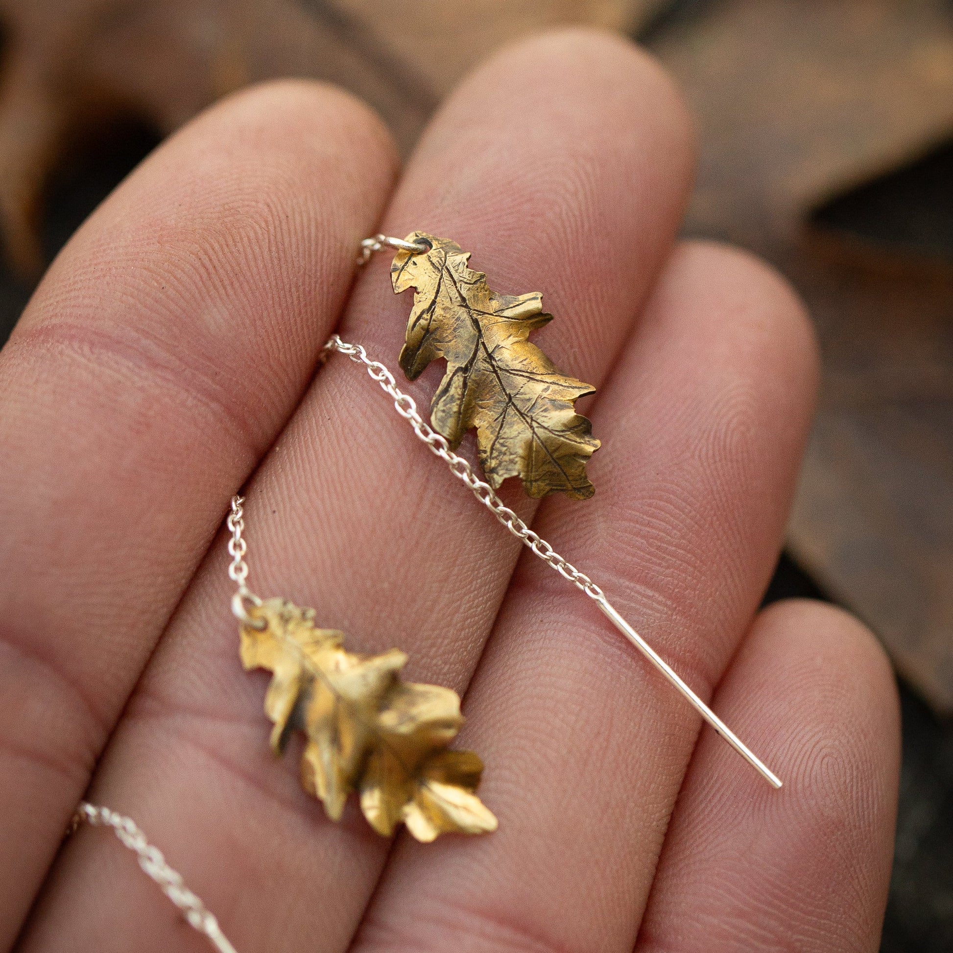 Brass gold colored oak leaf ear threader earring on a hand with a blurred natural background