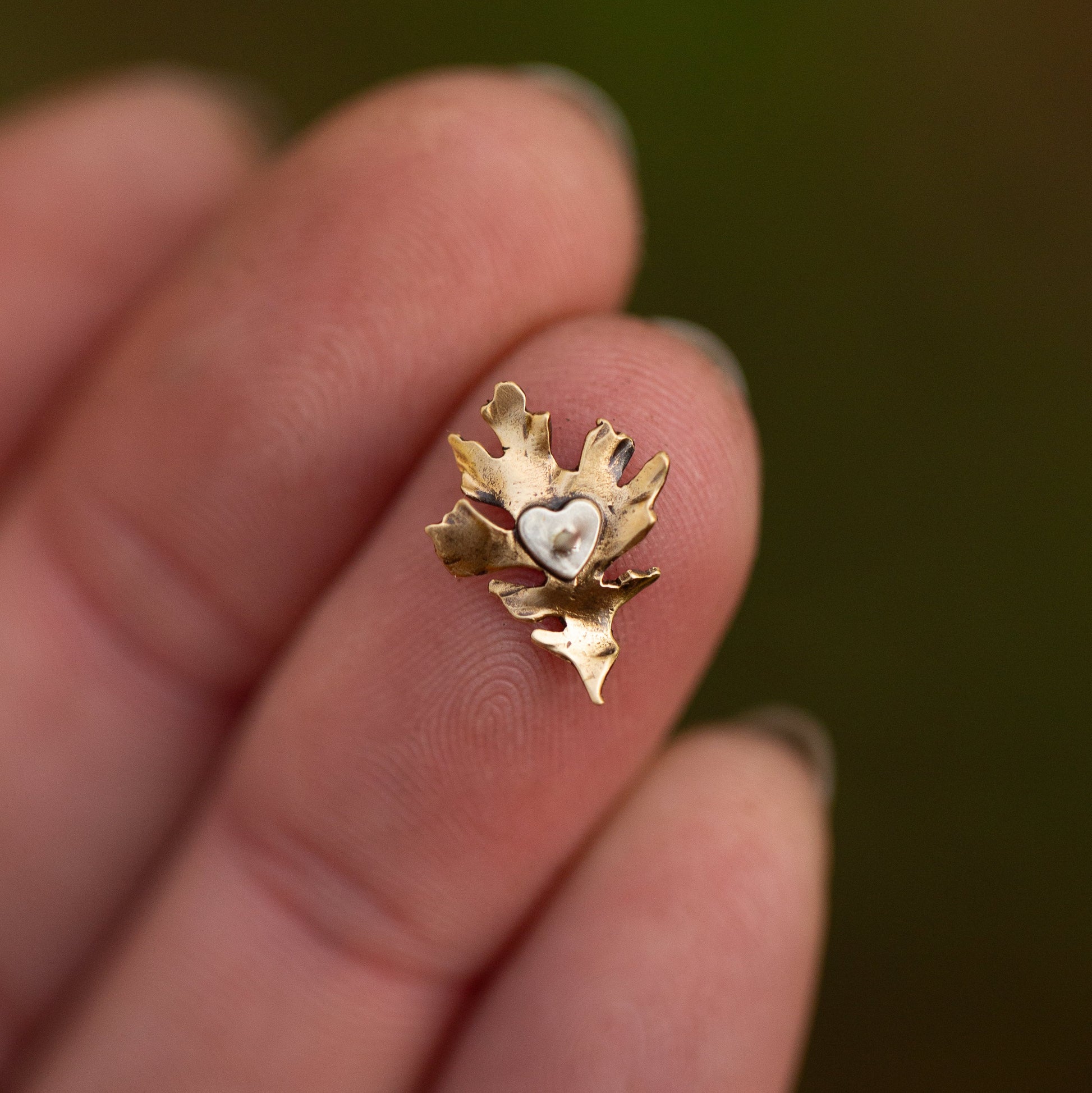 Gold heart-shaped earring held between fingers against a blurred background showing heart shaped silver back for an oak leaf stud earring