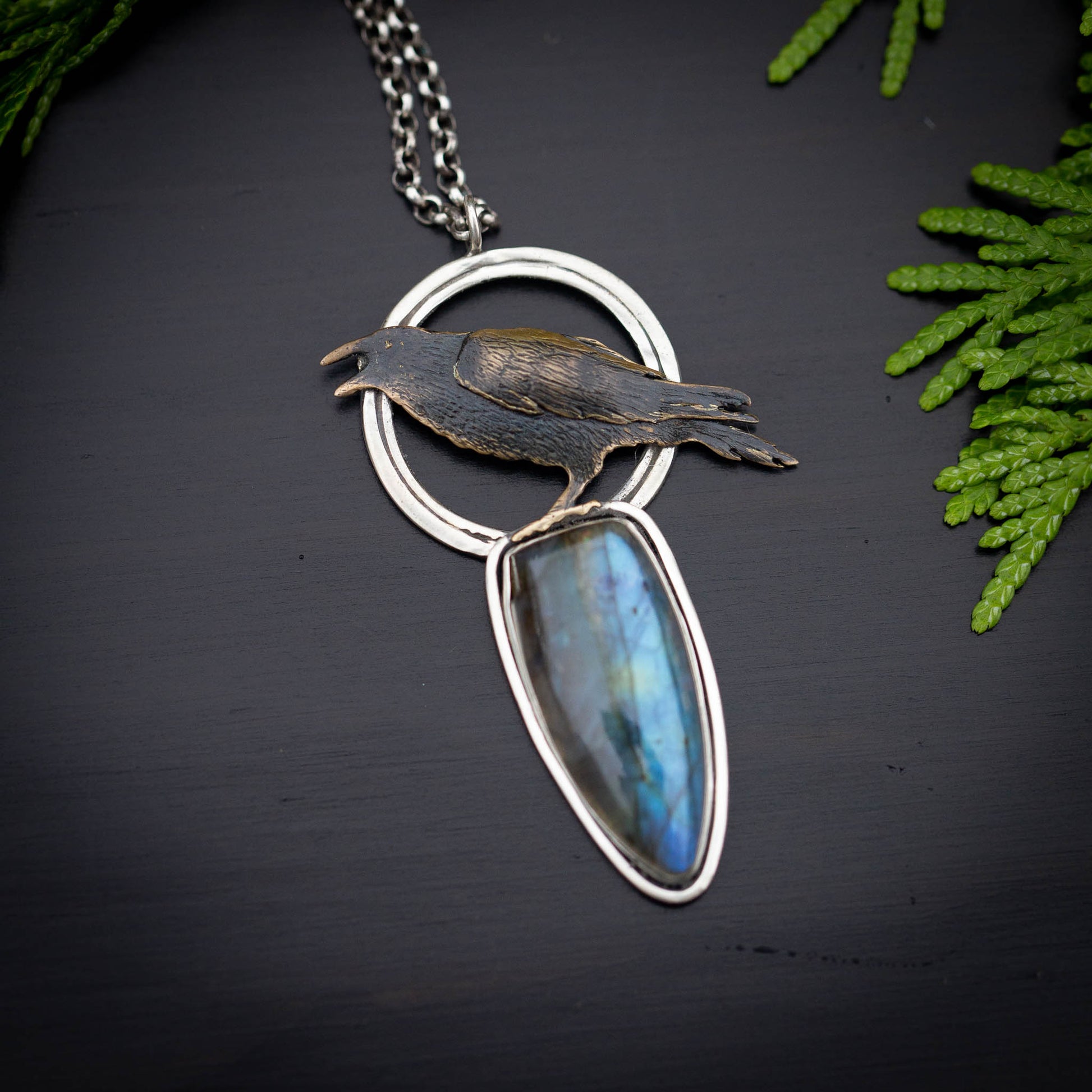 a necklace featuring a brass raven on an open silver circle utop a tear drop labradorite stone shown on a black background