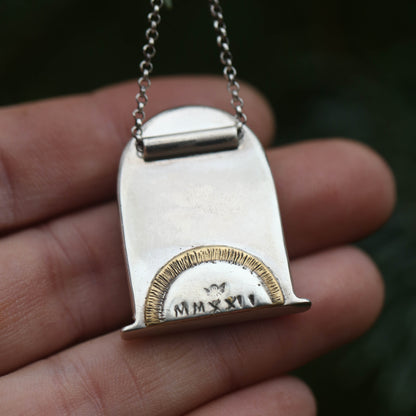 A hand showing the back of an arch shaped sterling silver and gold necklace with the date in roman numerals