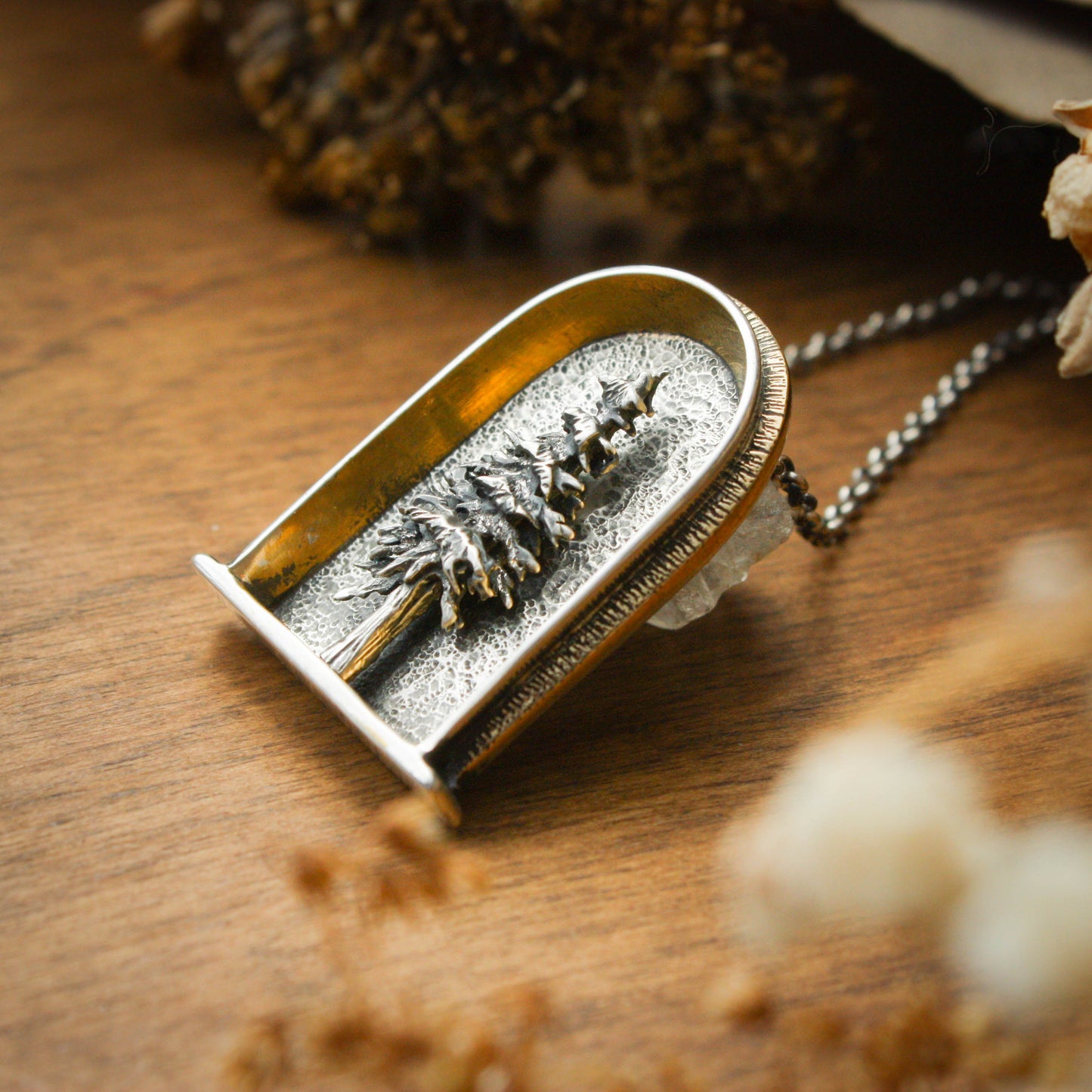 A sterling silver arched shrine shaped necklace featuring a three dimensional evergreen tree with a gold and brass border on a wooded background