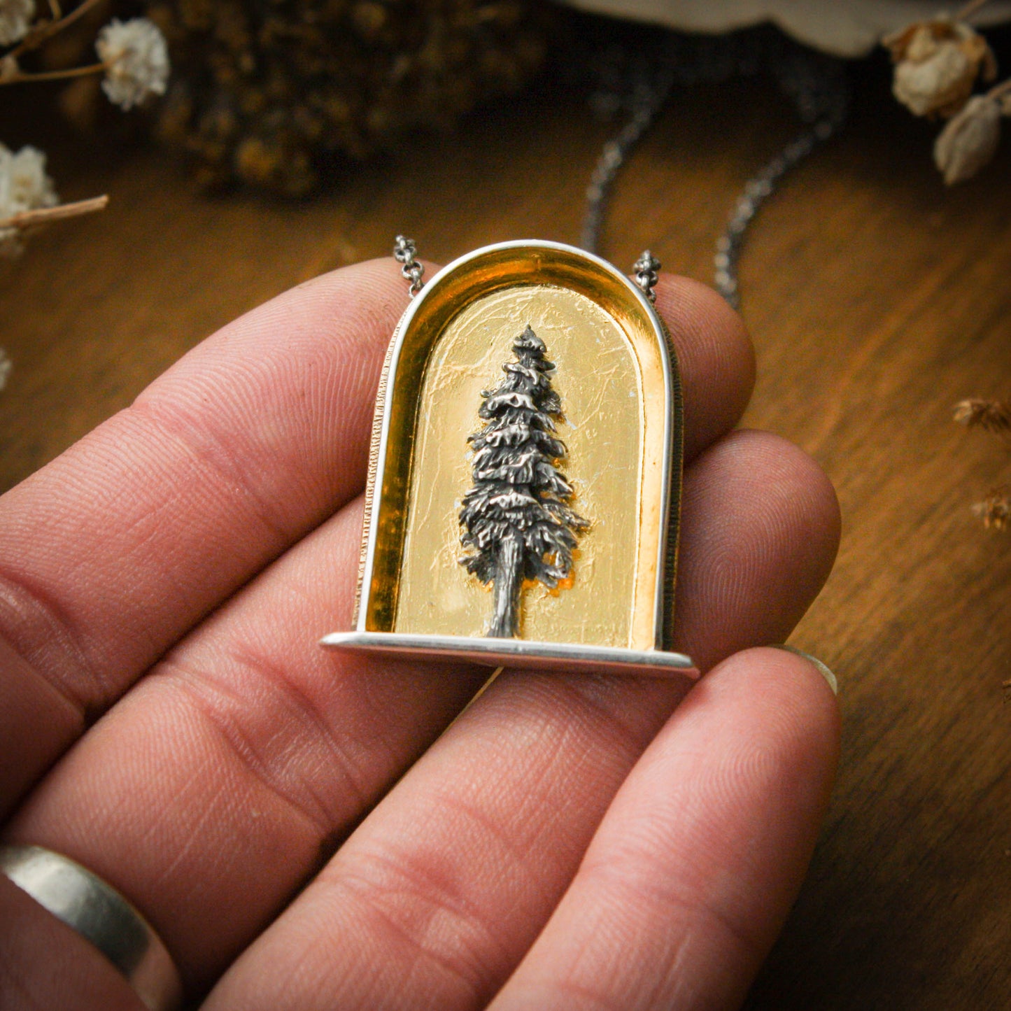 A hand with a sterling silver and 24k gold necklace in an arch shape featuring a layered sterlign silver redwood tree with a wood background